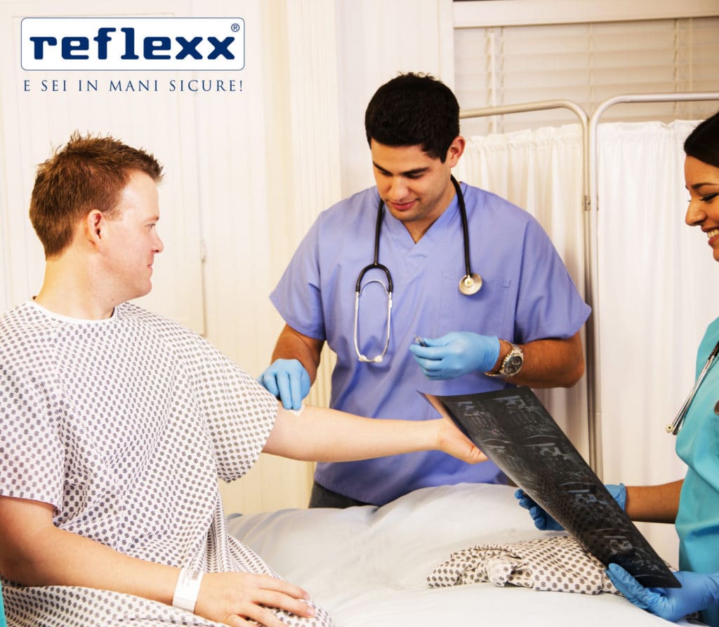 Hospital staff tend to patient in hospital room.  The mid-adult man is receiving an IV by nurse or doctor while female doctor or nurse reviews his recent x-ray results.  Other staff far left.  Staff wear scrubs and stethoscopes.